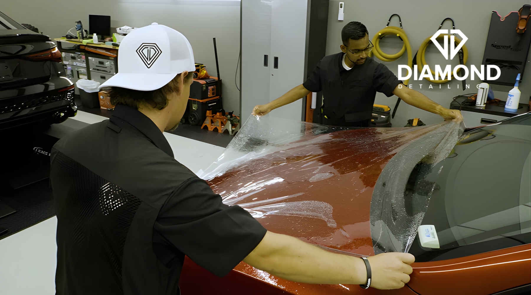 Close-up view of two men carefully stretching and smoothing Paint Protection Film over the hood of a red car, showcasing how What Is PPF looks in action during installation.