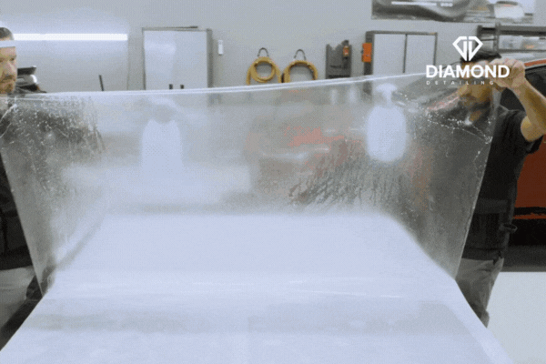 Two technicians at Diamond Detailing NC carefully stretch and position a large sheet of Paint Protection Film in a professional, well-lit garage, illustrating the precision and care behind their expert PPF installations.