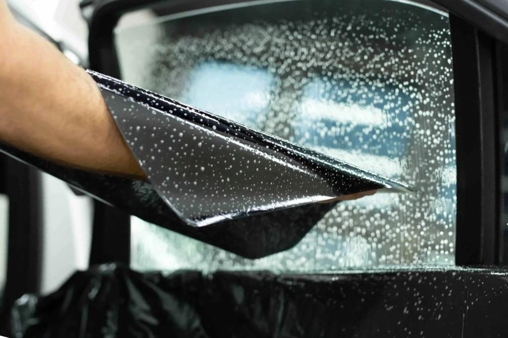 A close-up view of a professional applying dark window tint film to a car door window covered in water droplets, demonstrating precision during installation at Diamond Detailing while choosing the best tint for local needs in Troutman, NC.
