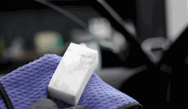Close-up of a white detailing sponge held with a blue microfiber towel, both used in interior auto detailing for precise cleaning of car surfaces.