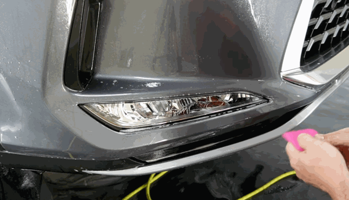 Close-up of a person using a pink squeegee to apply paint protection film to the front bumper of a silver SUV.